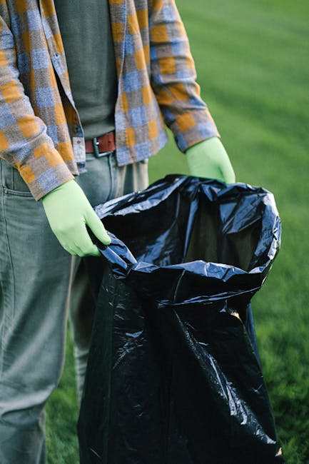 The image shows a person wearing a checkered shirt in shades of brown, beige, and gray, along with light green gloves, standing outdoors on a grassy area. They are holding open a large black rubbish bag made of plastic, which appears to be clean and ready for waste disposal. The person's torso and hands are visible, with the bag positioned in the foreground and their waist and part of their legs in focus. The background features a well-maintained green lawn, suggesting an outdoor setting, possibly a park or garden. The scene reflects an activity related to waste collection or disposal, aligning with private rubbish removal or on-site clearance services offered by companies like Rubbish Collection Bermondsey, providing an alternative to council-led rubbish collection. The lighting indicates natural daylight, emphasizing clarity and detail in the materials and textures of the clothing and rubbish bag, supporting the visual context of waste management and rubbish removal services.