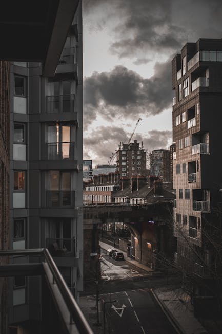 The image depicts a view from a balcony in an urban residential area during cloudy weather, with tall modern apartment buildings on either side of the scene. The building on the left features large glass windows, some illuminated from inside, with a smooth, light-colored exterior surface, while the building on the right shows a darker, textured facade with numerous balconies and windows. In the foreground, a metal railing runs along the balcony edge. Below, a narrow street with marked lanes and a few parked cars is visible, leading underneath an elevated motorway or railway bridge that spans across the scene. The background reveals a cluster of mid-rise and high-rise buildings, some under construction with visible cranes, set against a sky filled with dark, heavy clouds, indicating overcast weather. The overall environment suggests a dense, contemporary cityscape typical of an inner urban area, aligning with a context of private or independent waste management services that handle rubbish collection in such settings, as provided by Rubbish Collection Bermondsey.