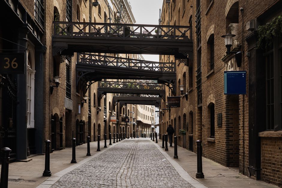 A narrow urban street paved with cobblestones, flanked by multi-storey red-brick buildings with black metal fire escapes and decorative street lamps mounted on the walls. Overhead, several dark metal pedestrian bridges with intricate latticework span between the buildings, creating a layered effect along the alleyway. The scene appears to be during daylight with natural light illuminating the facades, and a few pedestrians walking along the pavement. On the right, a blue rectangular sign is mounted on the building, with no visible text. The street is lined with evenly spaced black bollards, separating the pedestrian walkway from potential vehicle access, and the overall setting has a historic and industrial feel, typical of central city districts. This environment reflects a typical area where independent waste collection services, such as those offered by Rubbish Collection Bermondsey, might operate in urban residential or commercial zones, supporting alternative waste handling approaches in busy city areas.