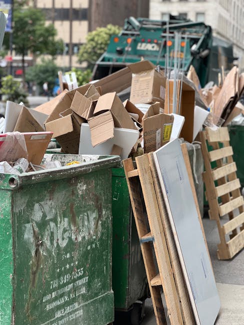 A large green waste skip filled with a variety of discarded cardboard boxes, some flattened and others still in their original shapes, alongside wooden pallets leaning against the side of the skip. The cardboard materials include both plain brown and white boxes, some with printed labels or markings visible. In the background, there is a partially visible green waste collection truck parked on an urban street, surrounded by a mixture of buildings, trees, and parked cars. The scene captures outdoor rubbish removal activity consistent with private waste collection services, with natural daylight illuminating the scene and highlighting the textures of the cardboard, wood, and metal surfaces. The arranged waste materials suggest an on-site clearance or bulk disposal typical of independent rubbish removal operations undertaken by companies such as Rubbish Collection Bermondsey, serving local commercial or residential clients in the area.