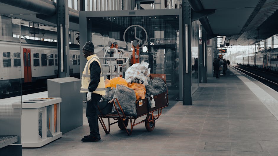 A man wearing a black beanie, dark clothing, and a reflective vest stands on a train station platform beside a red metal trolley loaded with various types of rubbish, including large black plastic bags, orange and yellow plastic bags, and miscellaneous discarded items. The trolley is positioned close to a waste receptacle, with the rubbish spilling over its sides. In the background, there are modern station features such as glass panels, metal structures, and multiple train tracks, with a train visible on the left side and another on the right, suggesting an ongoing or recent waste collection process related to station cleanliness or private rubbish handling services like those offered by Rubbish Collection Bermondsey. The overall environment appears to be a busy, urban transportation hub with natural lighting illuminating the concrete flooring and the surrounding infrastructure.