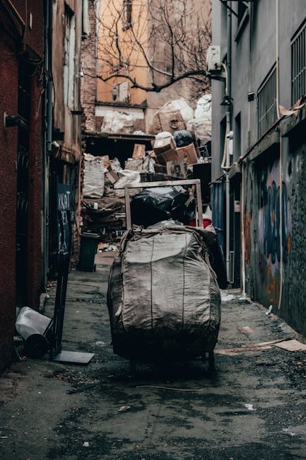 An alleyway with a narrow paved pathway flanked by tall, weathered buildings on both sides. In the center foreground, a large wheeled rubbish bag, made of gray, slightly crinkled material, is positioned upright, possibly indicating recent collection or disposal activity. Behind it, a cluttered pile of mixed waste materials, including cardboard boxes, plastic bags, and other debris, is stacked against a wall at the end of the alley. The alley appears somewhat confined and cluttered, with scattered small debris resting on the ground. The walls on either side are adorned with graffiti and various markings, while tree branches are visible overhead, casting faint shadows and adding an organic element to the scene. The environment suggests a private waste collection area, highlighting the need for efficient rubbish removal services, such as those offered by Rubbish Collection Bermondsey, for on-site clearance or alternative waste handling in urban settings.